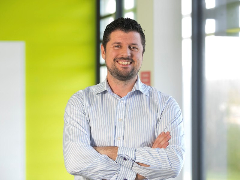 Man in striped shirt with arms folded standing in front of a green wall and a window.