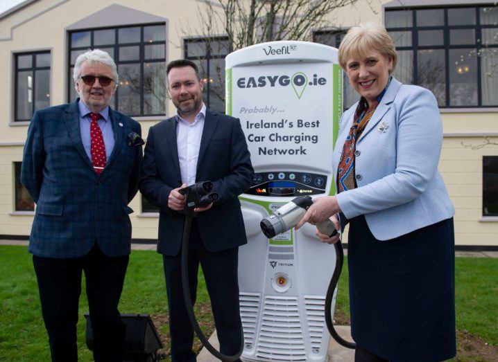 Two men in suits and a woman in a light blue blazer holding EV chargers at an EasyGo charging station.