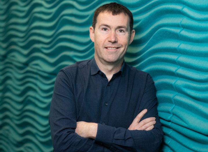 Man in navy shirt and black hair stands with arms folded against a green wavy wall.