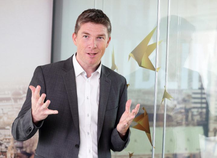 A man in a shirt and blazer gestures as he speaks next to a glass wall decorated with illustrations of golden birds.