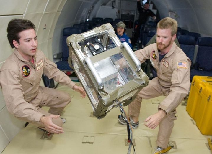 Two male researchers in beige astronaut outfits looking at an experiment in zero gravity.