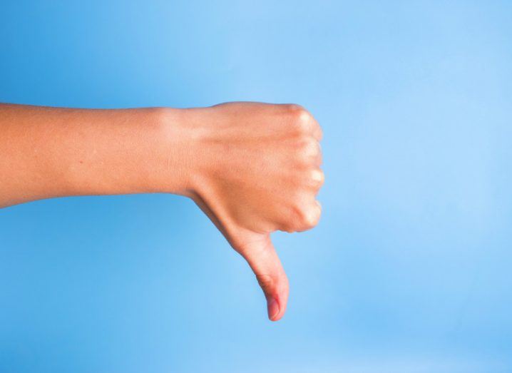 Close-up of woman's hand gesturing thumbs down against blue background, indicating another bad scandal at Facebook.