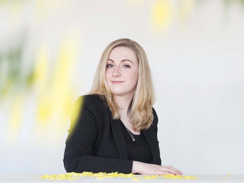 Woman with blonde hair and black jacket sits with one arm on a table strewn with flower petals.