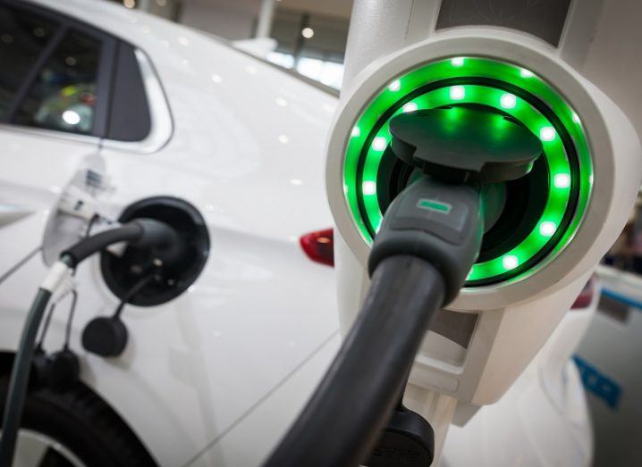 Close-up of an EV charging point lit up with green lights with a white car charging behind it.