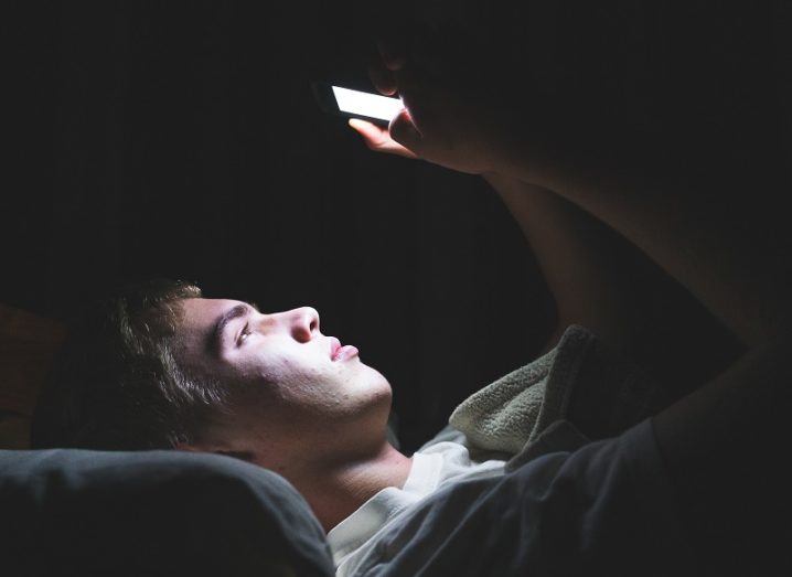 A teenage boy lying in bed in the dark with his phone screen lighting up his face.
