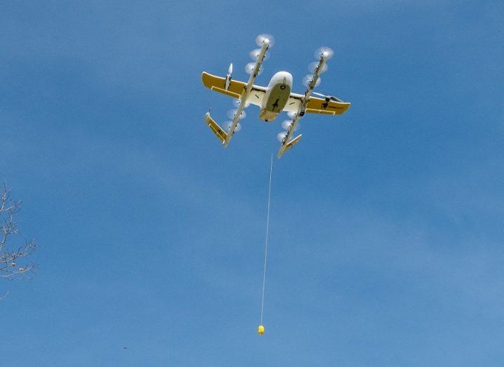 The Wing drone hovering over a delivery site with its tether unfurled against a blue sky.
