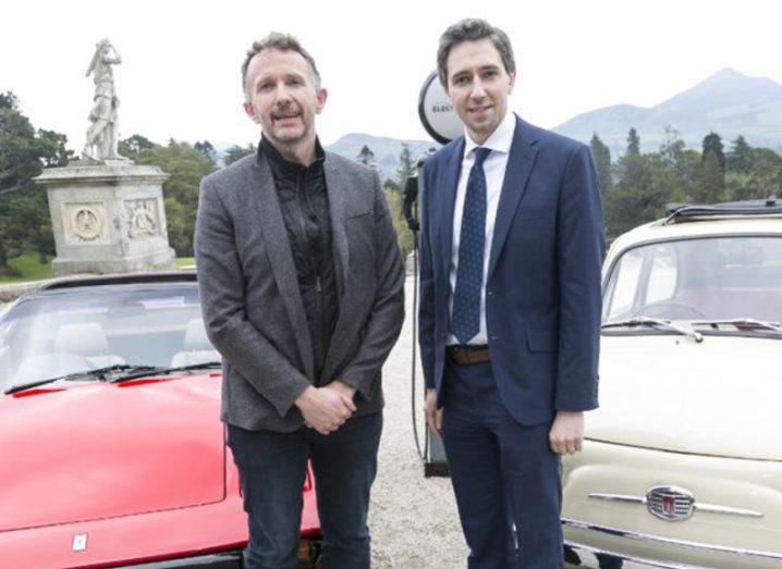 Two men in suits stand in front of a pair of classic cars.