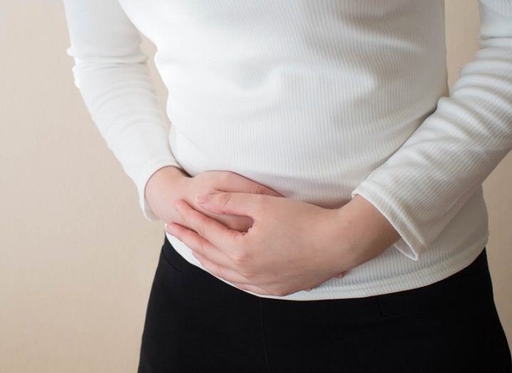 Woman in a white jumper and black trousers holding her stomach in pain, symbolising endometriosis.