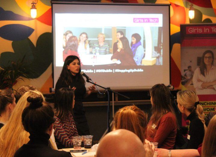dark-haired woman speaking into mic in front of an audience with a Girls In Tech presentation behind her.