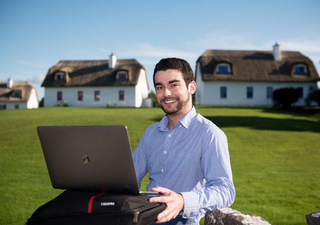 Man in blue shirt sits with a laptop in front of Irish cottages.
