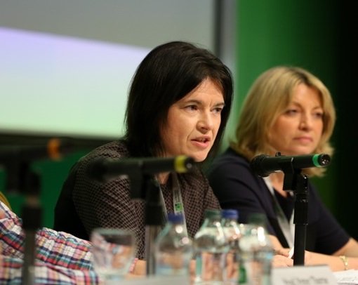 Áine Ryall speaking into a microphone on stage at the Citizens' Assembly with a projector screen in the background.