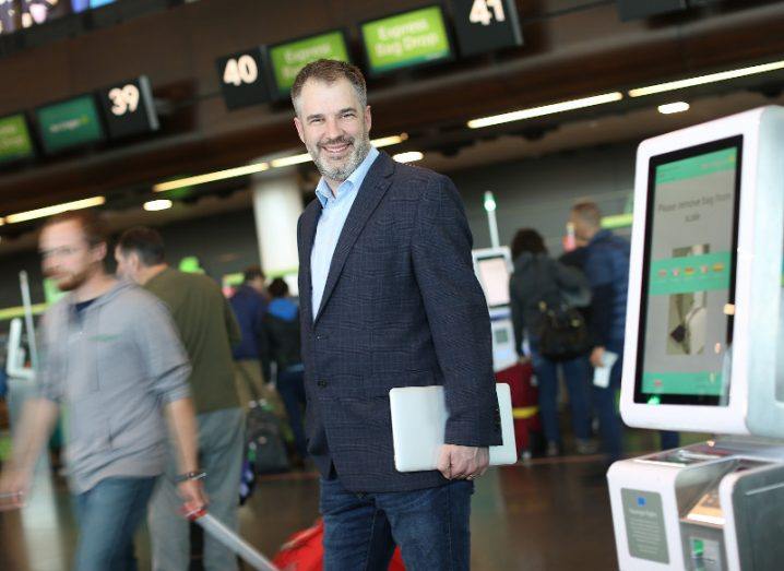 Man in dark jacket standing in departures area of Dublin airport holding a tablet computer.