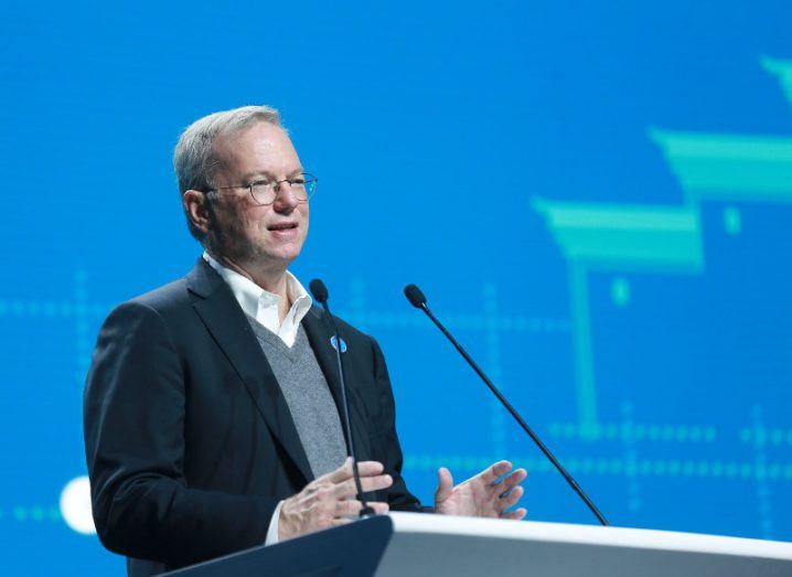 Middle-aged man in glasses and suit delivers speech at a podium. against light blue background.