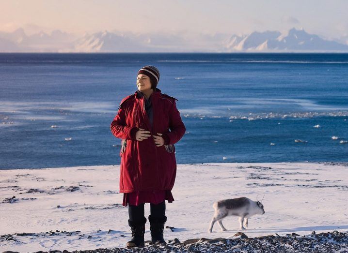 Distant shot of Jenny Hanafin in a long red coat standing against an icy coast off Antarctica.