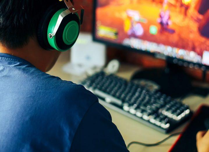 A teenager with a blue t-shirt with green headphones playing a video game on a PC.
