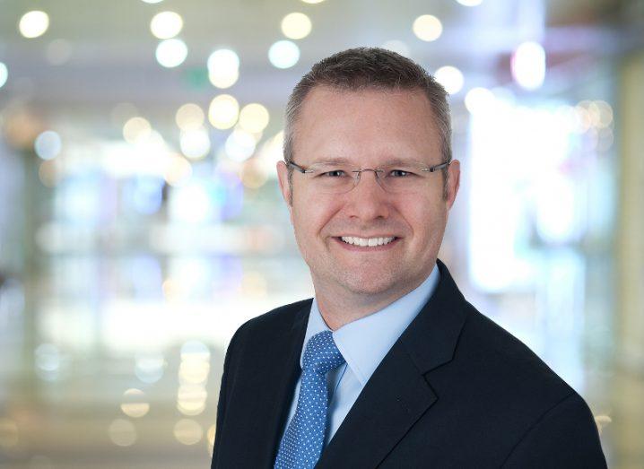 suited man with frameless glasses smiling at the camera against a blurred out office background.