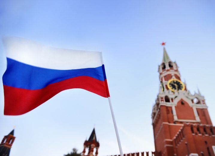View of the flag of Russia billowing in the wind against a clear sky with view of building in distance.