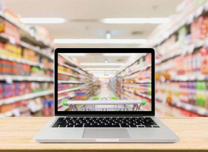 An open laptop on a table in front of a long aisle of products for sale. The laptop screen duplicates the view of the aisle.
