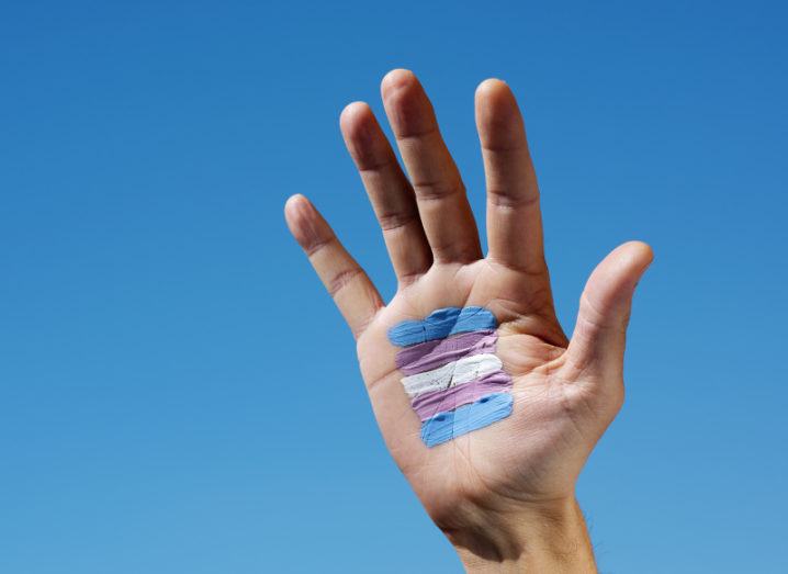 A hand is held up against a blue sky. Painted on the palm are the blue, pink and white stripes of the trans pride flag.