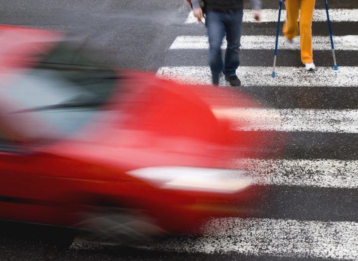 Blurred image of a red car driving through a pedestrian crossing with two people already crossing.