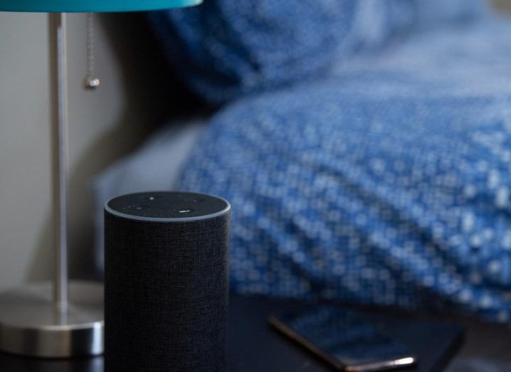 An Amazon Echo smart speaker on a bedside table beside a bed with a blue and white spotted duvet.