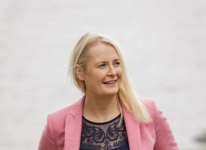 close-up of blonde woman wearing light pink blazer standing outside against a seaside background.