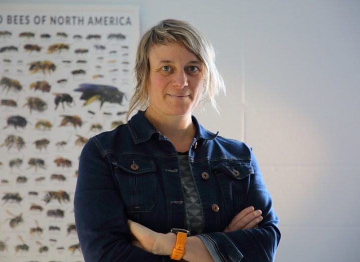 Dr Ilona Naujokaitis-Lewis in a denim jacket crossing her arms and smiling with a bee species chart behind her.