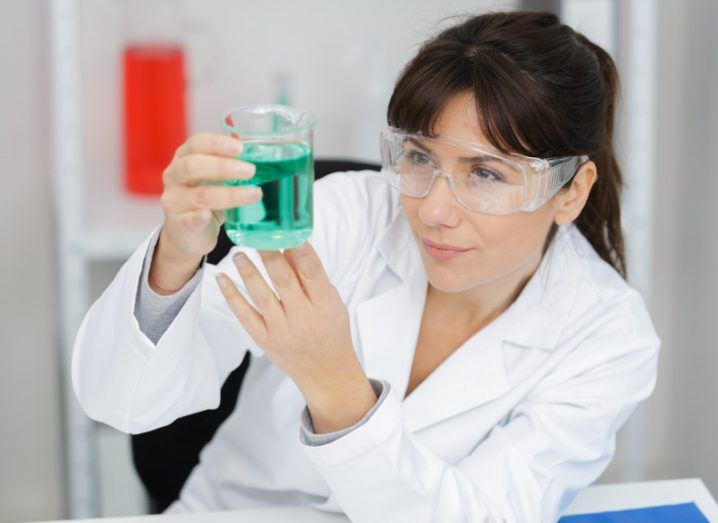 Female scientist in white coat holding a vial of green liquid.
