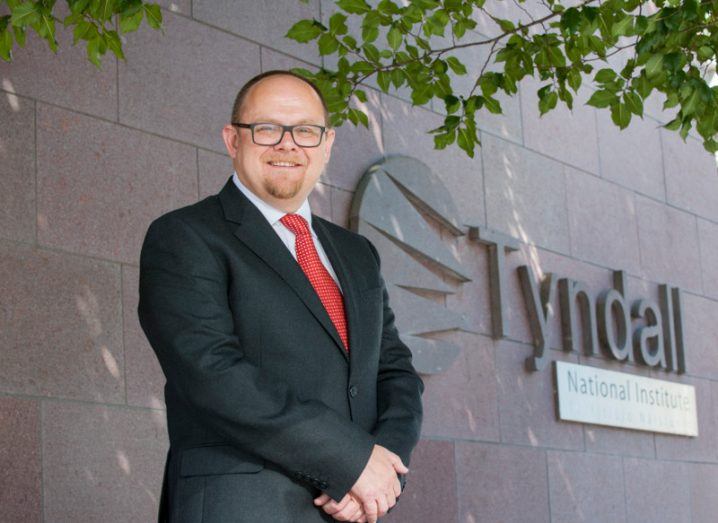 middle-aged man with glasses wearing a suit and standing outside beside a stone wall with a sign for Tyndall National Institute.