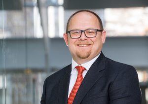 close-up of middle-aged man with glasses and stubble wearing a suit and red tie, standing outside.