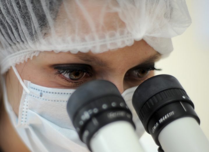 A scientist wearing a hairnet and medical mask looks into a microscope.