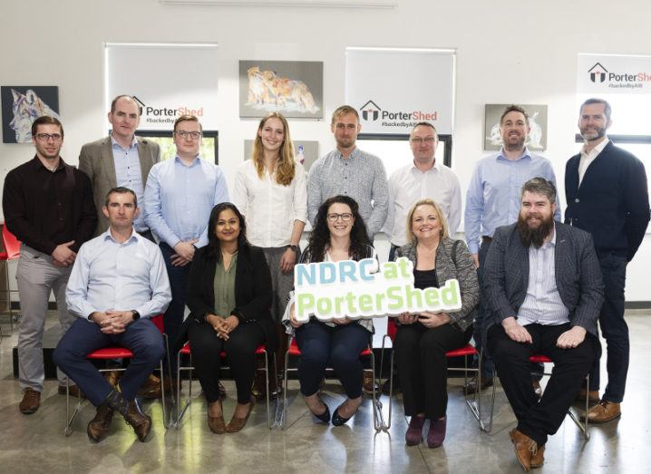 A row of eight people stand behind a row of five seated people. A woman seated in the centre holds an NDRC at PorterShed sign.