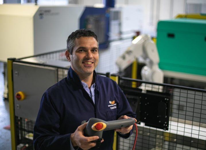 Dr Declan Devine smiling in blue overalls holding a piece of equipment in a factory setting.
