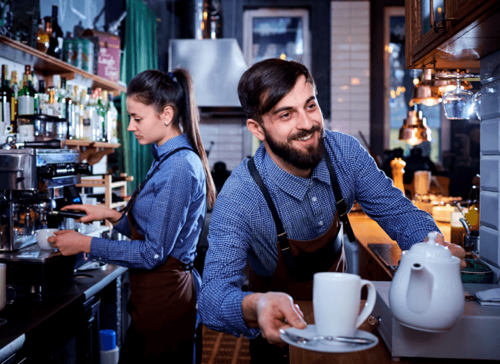 Two waiters wearing blue shirts and red aprons work in a busy kitchen. One is making a cup of coffee, while the other is holding a teapot and a teacup.