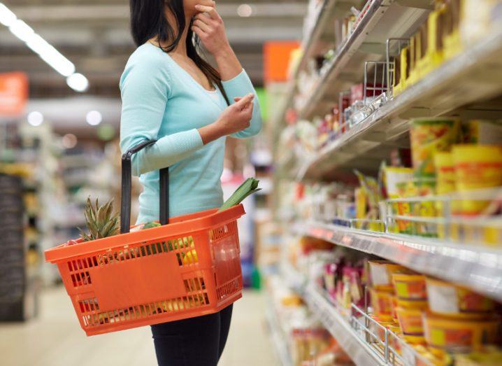 Woman in grocery aisle with orange shopping basket dangling off arm with hand on chin mulling prospective purchases.