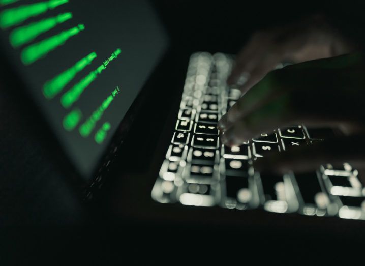 View of hands floating over the illuminated keys of a laptop, the screen of which is black with green back-end coding interface text.