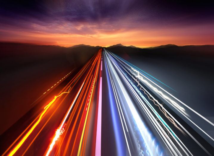 Fast neon-coloured traffic light trails on a highway stretching into a darkening sunset horizon.