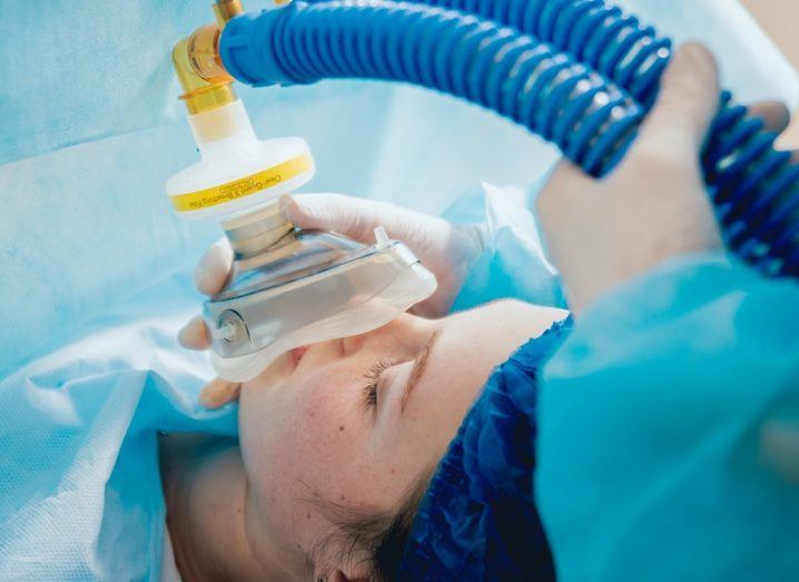 A patient covered in a blue sheet is prepared for surgery as a doctor places an anesthesia machine over her face.