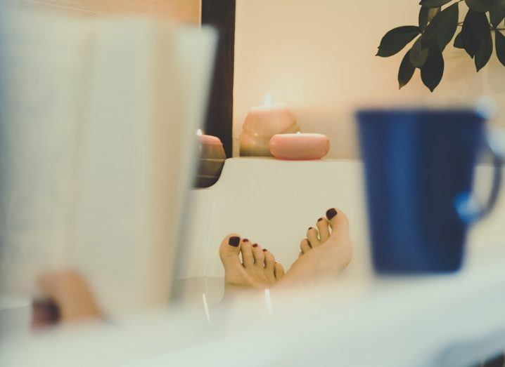 Shot behind a person lying in a bath with their feet out of the water, reading a book with a coffee cup beside them.