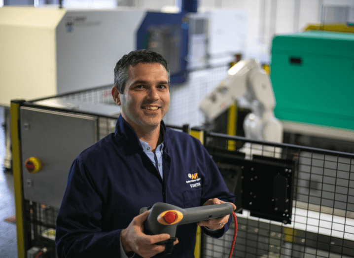 Declan Devine from AIT holds a grey device while standing in front of the polymer testbed, which is guarded by a railing. He has dark hair and is wearing a navy jumpsuit.