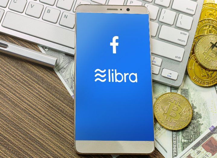 A smartphone bearing the Facebook Libra currency logo sits on top of a keyboard with some gold 'bitcoins' nearby.