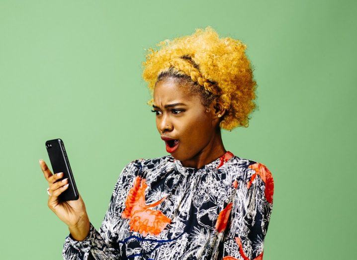 Woman with yellow hair wearing a vibrant black, white and orange shirt making a shocked face while looking at a smartphone against a green backdrop.