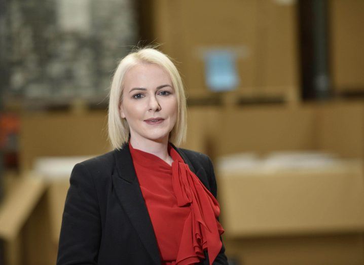 A woman with blonde hair wearing a black suit and red shirt stands in an office.