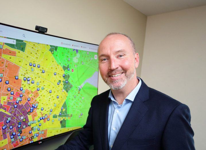 man with grey beard wearing blue shirt and navy blazer smiles broadly while standing next to a screen with geolocation information in red, yellow and green.