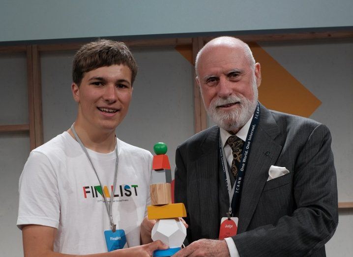 Fionn Ferreira, a young man in a white T-shirt, and Vint Cerf, an older man in a suit, holding the Google Science Fair trophy following the award win.
