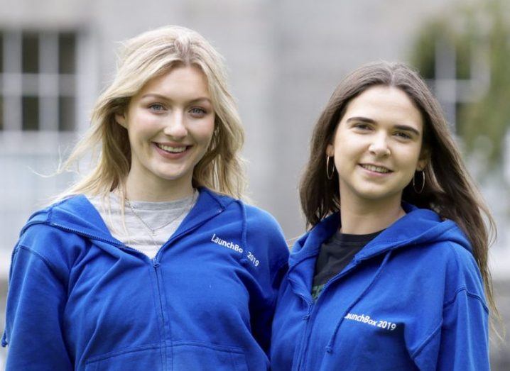 two young women, one blonde and one brunette, standing outside wearing blue hoodies branded with LaunchBox logo.