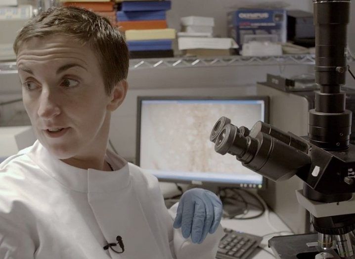 Dr Eilís Dowd in a white lab outfit looking over her shoulder in front of a microscope and computer with data on screen.