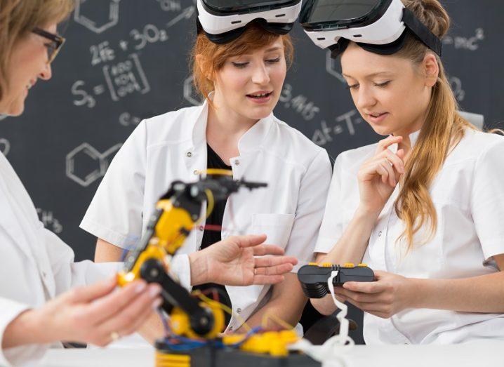 Two students in white lab coats wearing VR headsets while a lecturer tinkers with a robotic arm against a chalkboard background.