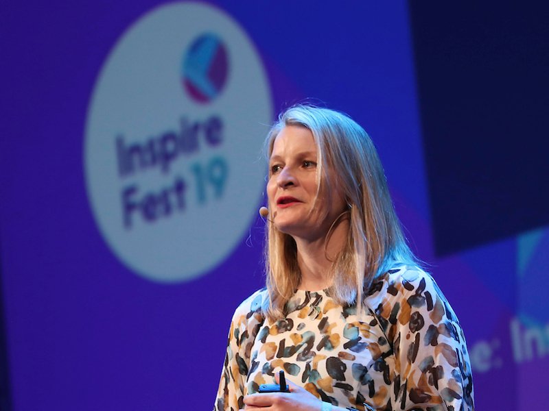 Woman with long blonde hair wearing head mic and patterned dress speaking to crowd at Inspirefest 2019 in front of purple backdrop.