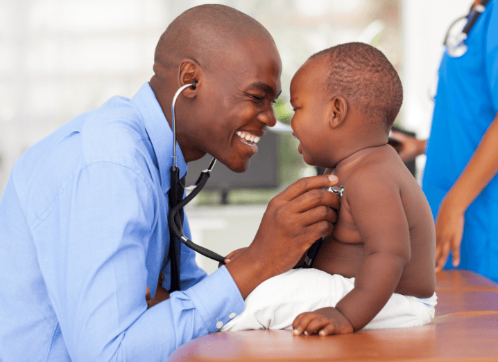 A doctor smiling warmly as he measures a baby's heartbeat with a stethoscope.